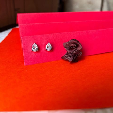A pair of silver teardrop stud earrings with clear stones displayed on a bright pink card next to a dark brown satin rose, set against a textured orange-red surface.