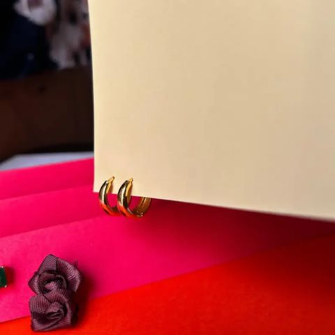 A close-up of three small gold hoop earrings resting against a cream-colored surface, with bright pink and red paper layers and a small fabric rose in the foreground.