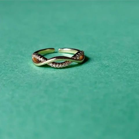 A twisted open ring with a silver-tone metal band, featuring a row of small clear stones on one side and a curved amber-brown inlay on the other, set against a solid teal-green background.