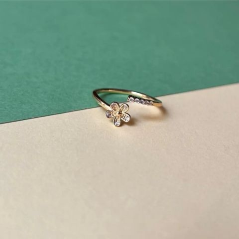 A delicate gold open-band ring featuring a small flower charm with clear stones and a row of tiny pavé-set clear stones, resting on a split green and beige surface.
