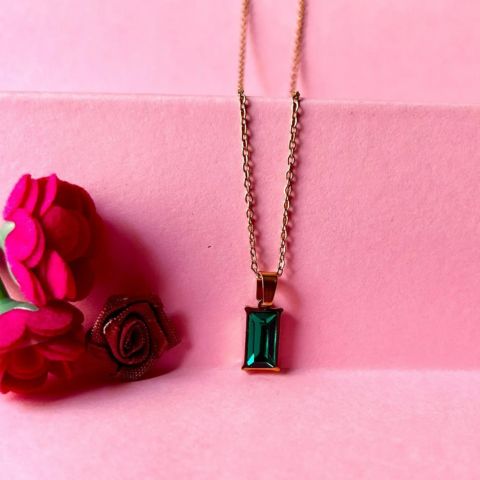 A rose gold chain necklace with a rectangular green emerald-cut gemstone pendant, displayed on a pink background next to red fabric roses.