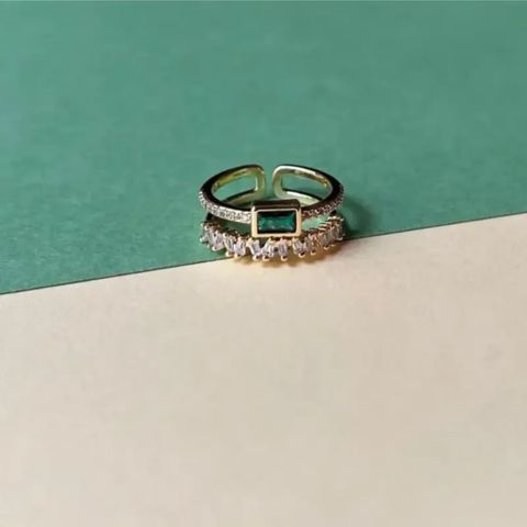 A gold-plated open band ring with a central rectangular green stone and rows of clear stones, photographed on a split green and cream background.