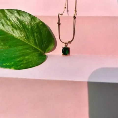 A gold necklace with a dark green oval pendant resting on a pink step, next to a large green leaf.