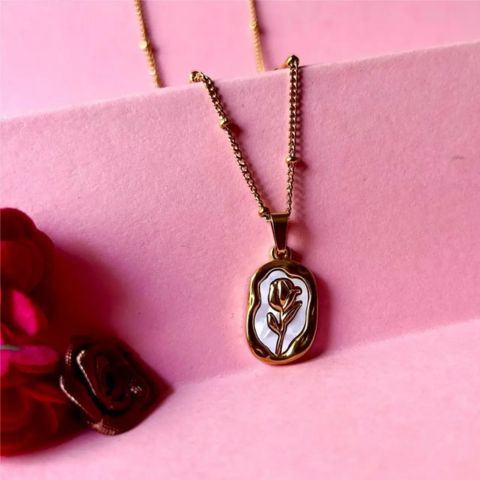 A gold-tone necklace with an irregular oval pendant featuring a white inlay and a rose motif, displayed on a pink background with a pink card and dark red rose props.