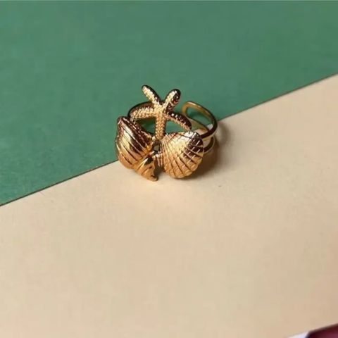A gold-toned ring featuring a textured starfish design wrapping around two seashells, displayed on a split green and beige background.