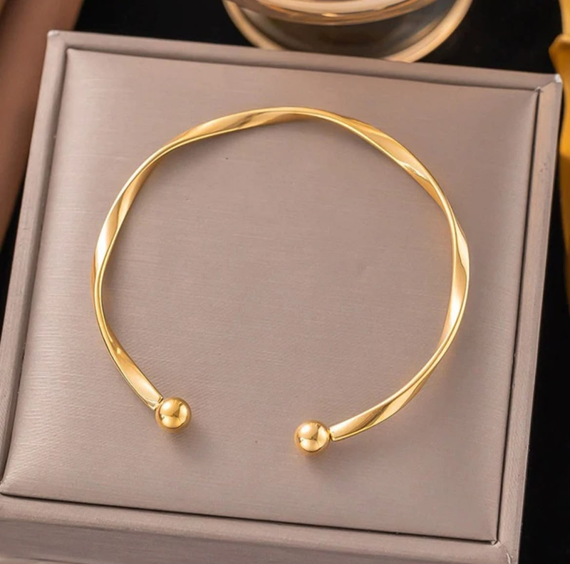 A twisted open cuff bracelet in polished yellow gold, displayed on a textured gray jewelry box.