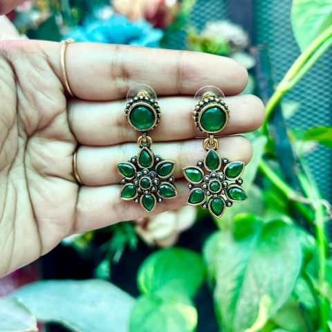 A pair of green stone drop earrings with ornate gold-toned settings, held on a person's hand against a blurred background of green leaves and flowers.
