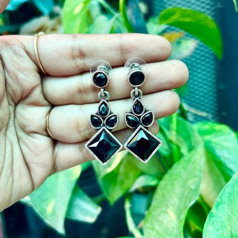 A pair of silver-toned dangle earrings with black faceted gemstones, held in a person's hand against a background of green houseplants.