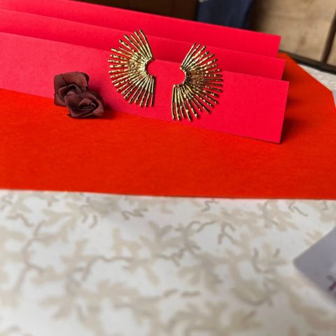 A pair of gold-toned fan-shaped earrings displayed on red card holders, with a small dark red fabric rose beside them on a red surface.