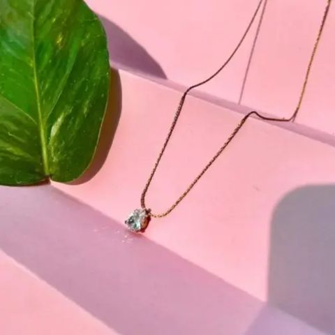 A delicate gold chain necklace with a small heart-shaped clear gemstone pendant, styled on a pink geometric surface next to a green leaf.
