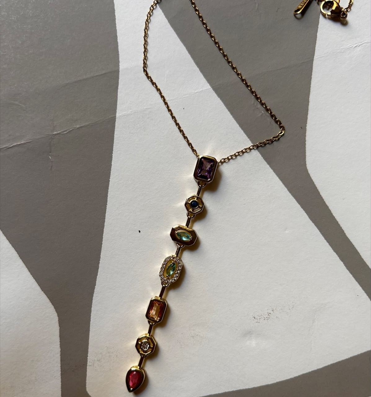 A gold-toned lariat-style necklace with a long drop chain featuring multiple colorful gemstones in different shapes and colors, laid out on a white and gray geometric background.