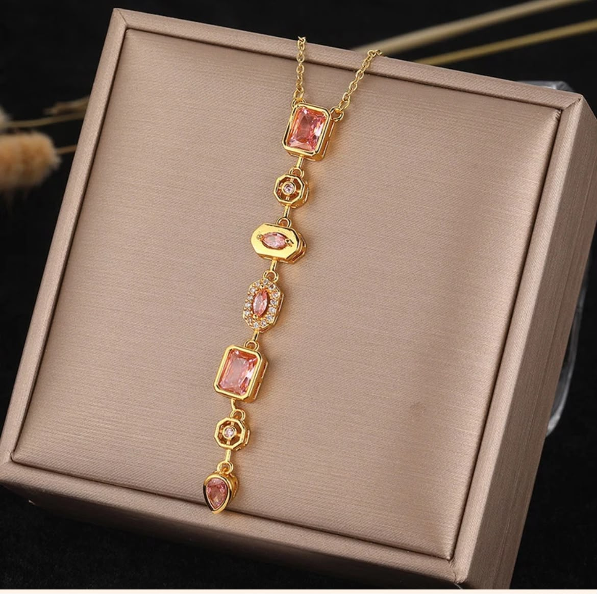 A gold-plated chain necklace featuring multiple pink gemstone charms in rectangular, heart, and oval shapes, displayed on a champagne-colored satin jewelry box.