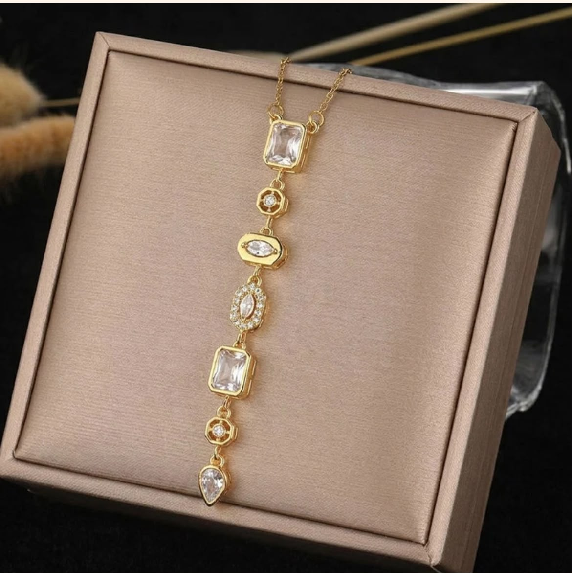 A gold-tone drop necklace with clear rectangular and oval gemstones, displayed on a champagne-colored satin jewelry box.